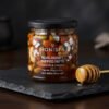 A glass jar of Honista Real Honey Dipped Nuts with almonds cashews and edible silver varq leaf on a black slate slab with golden wooden honey dipper.