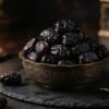 Exquisite dark VIP Royal Ajwa Al-Madinah dates piled into an ornate engraved brass bowl on dark slate with soft warm candlelight reflection, ultra-premium gourmet e-commerce food photography