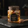 Hexagonal artisan honey jar filled with whole cashews, almonds, and pistachios submerged in golden translucent mountain honey with edible silver leaf accents on a black slate slab with a wooden honey dipper, luxury gourmet confection.