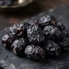 Macro close-up portrait of dark authentic Saudi Ajwa dates with natural wrinkles and deep purplish-black luster on dark royal stone plate.